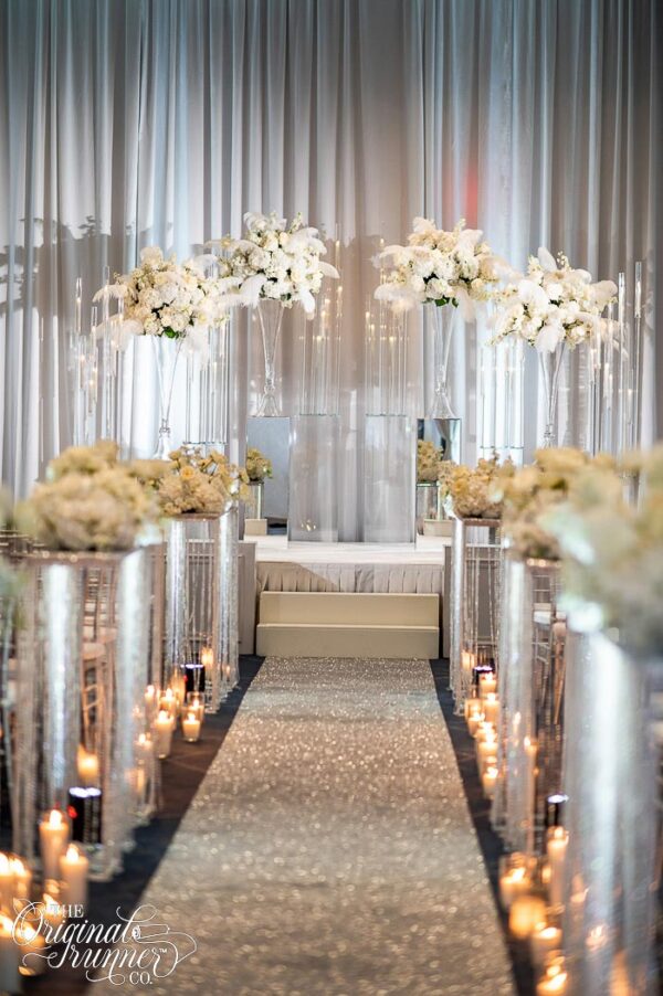 The JW Marriott in Nashville, Tennessee, provided the setting for an elegant affair celebrating the celebrity wedding of Tiffany Roy to retired football player Michael Oher, who was the inspiration for The Blind Side movie featuring Sandra Bullock. The ballroom was transformed into a stunning and sparkling space, creating a memorable experience for the couple and their guests. Candles lined the aisle, casting a warm and inviting glow throughout the room. The centerpiece of the decor was a silver glitter aisle runner, installed over the pattern ballroom carpet that sparkled beautifully in the candlelight. This added a touch of glamour and elegance to the event. At the head of the room, a raised platform served as the focal point for the ceremony. The platform was decorated with tall white floral displays featuring white feathers, adding a sense of sophistication and luxury. This chic decor enhanced the overall ambiance of the ballroom, making the wedding a truly memorable occasion for all those in attendance.