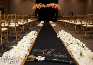 The Riverfront Banquet Center in Flint, MI, provides the perfect backdrop for this elegant wedding ceremony. The ballroom has been transformed into a space of beauty and romance. Gold delicate chairs set the stage for guests to take their seats and witness the exchange of vows. At the altar, a simple yet elegant arbor adorned with lush white florals creates a focal point for the ceremony. Plush white floral arrangements line the aisle, adding to the sense of luxury and sophistication. Central to the aisle is a stunning black aisle runner, adorned with a delicate gold border along the edges. Personalized with the names "Damen & Nafisah" in thin white cursive script, and featuring a hand-painted gold "F" to represent their new last name, the runner adds a touch of elegance and individuality to the space. Their wedding date is painted underneath, serving as a beautiful reminder of this joyous occasion. In this photo, the couple has placed their "Broom" as part of their "jumping the broom" ceremony, a tradition steeped in the history of the Black community. This ritual symbolizes the couple's commitment to each other and their journey into marriage, adding depth and meaning to their wedding celebration.