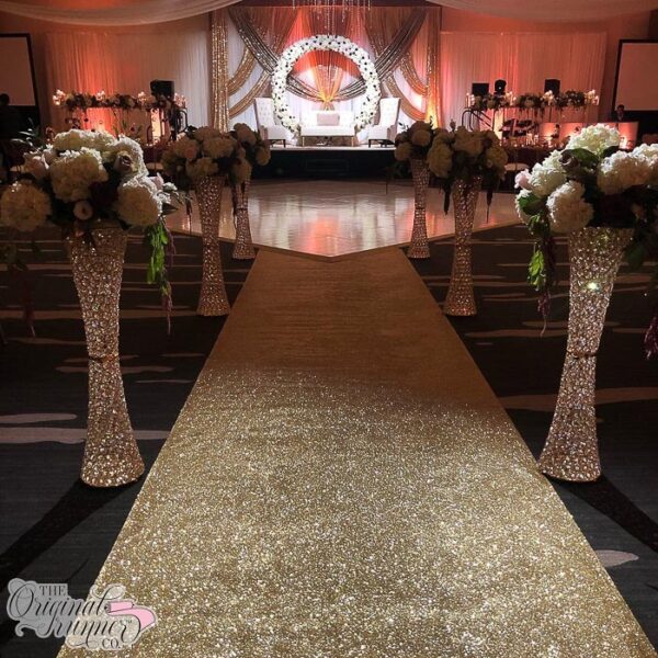 The Hyatt Regency Wichita in Kansas sets the stage for a captivating Indian wedding in its elegant ballroom, adorned with moody lighting that adds a touch of allure to the atmosphere. At the forefront of the room stands a magnificent white floral circle-shaped arbor, illuminated by spotlights to ensure it stands out as a focal point for the wedding party and guests. Attention is drawn to the aisle, which exudes sophistication and glamour. A stunning gold glitter aisle runner adorns the pathway, its shimmering surface perfectly illuminated to create a magical effect. Flanking the aisle are tall crystal pedestals, each adorned with lush white hydrangea floral bouquets, adding an element of opulence and refinement to the scene. This setting is truly elevated and elegant, embodying the essence of a sophisticated wedding celebration.