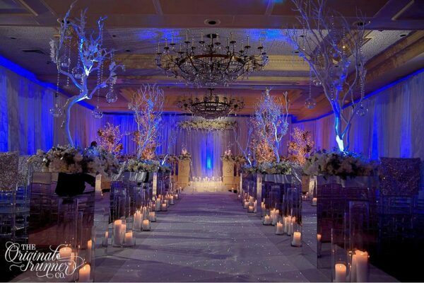 This wedding epitomizes elegance, with the ballroom transformed into a breathtaking winter wonderland. The grandeur of the room is enhanced by its architectural details, including large chandeliers and recessed ceilings, which create a sense of opulence. To add a soft and intimate touch, sheer drapery envelops the room, creating a dreamy ambiance. Moody blue uplighting further enhances the atmosphere. As guests make their way down the aisle, they are greeted by mirrored floral pedestals that reflect the flickering light of rows of candles and tall white tree branches adorned with hanging crystal details. However, the true focal point is the iridescent white aisle runner, which seems to shimmer and dance in the light, creating a magical and unforgettable experience for all who witness it.