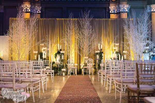 Set in the enchanting atmosphere of The Weylin in Brooklyn, NY, this wedding transports guests to a magical winter wonderland. The theme, evoking the serene beauty of the season, is brought to life through carefully curated decor elements. At the head of the room, frosted white tree branches reach skyward, creating an ethereal backdrop reminiscent of a snowy forest. Illuminated by the soft glow of candles, arranged in varying heights and interspersed with dark greenery, the display adds warmth and ambiance to the space. Against this backdrop, rows of white chairs blend seamlessly with the wintry theme, their simplicity enhancing the overall elegance of the setting. However, it's the centerpiece of the aisle that truly captivates attention—a rose gold glitter wedding runner that shimmers and sparkles under the soft, moody lighting. Each flicker of light accentuates the glittering surface, casting a spellbinding effect throughout the room and infusing the atmosphere with an enchanting allure.