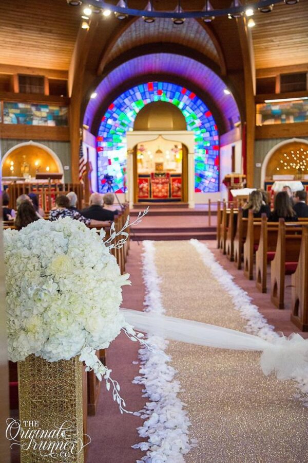 A stunning wedding at the Assyrian Orthodox Church of Virgin Mary in Paramus, New Jersey, creates a breathtaking scene for the couple's special day. The church features an exquisite arched altar with beautiful colorful glass mosaic details that radiate hues of blue, green, and purple, providing a mesmerizing backdrop. As guests enter the aisle, they are greeted by elegant golden pedestals adorned with lush white hydrangea floral bouquets. These opulent arrangements frame the entrance and set the stage for the rest of the ceremony. The aisle itself is a showstopper with a glittering champagne gold aisle runner that sparkles under the church's lighting. This stunning runner creates a luxurious pathway for the couple and their wedding party to walk down. To soften the look and enhance the elegance of the setting, lush white rose petals are carefully placed along the edges of the aisle runner, adding a romantic touch to the already enchanting space.