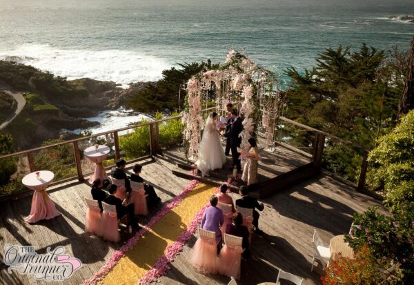 This intimate wedding took place at the Hyatt Carmel Highlands in Carmel, California, offering a stunning outdoor deck with breathtaking ocean views. Despite its petite size with under 10 guests, the ceremony did not lack grandeur or elegance. In the corner of the deck, a raised platform provided the perfect stage for the couple to recite their vows. The arbor was beautifully decorated with cascading soft pink florals and dripping with crystal strands which catch the light. The couple, a stunning Asian bride dressed in a traditional organza ballgown with lace sleeves, and the groom in a classic black suit, stood facing each other under the arbor as they exchanged vows. The chairs set in two rows were adorned with blush pink organza and rose details, enhancing the intimate and elegant atmosphere. For the aisle, the couple chose a stunning gold glitter runner that sparkled in the sunlight, adding a touch of glamour to the ceremony. Its edges were delicately decorated with light pink rose petals, harmonizing the decor and creating a cohesive and romantic look. This small wedding proved that even intimate ceremonies can be just as grand, with its elegant touches and picturesque setting making it an unforgettable experience for the couple and their guests.